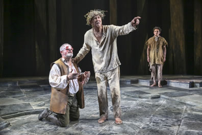 Gloucester in brown leather vest kneels as Lear in white pajamas and weed crown stands by with a hand on Gloucester's shoulders and the other hand pointing. Edgar in ragged clothes stands at the back.